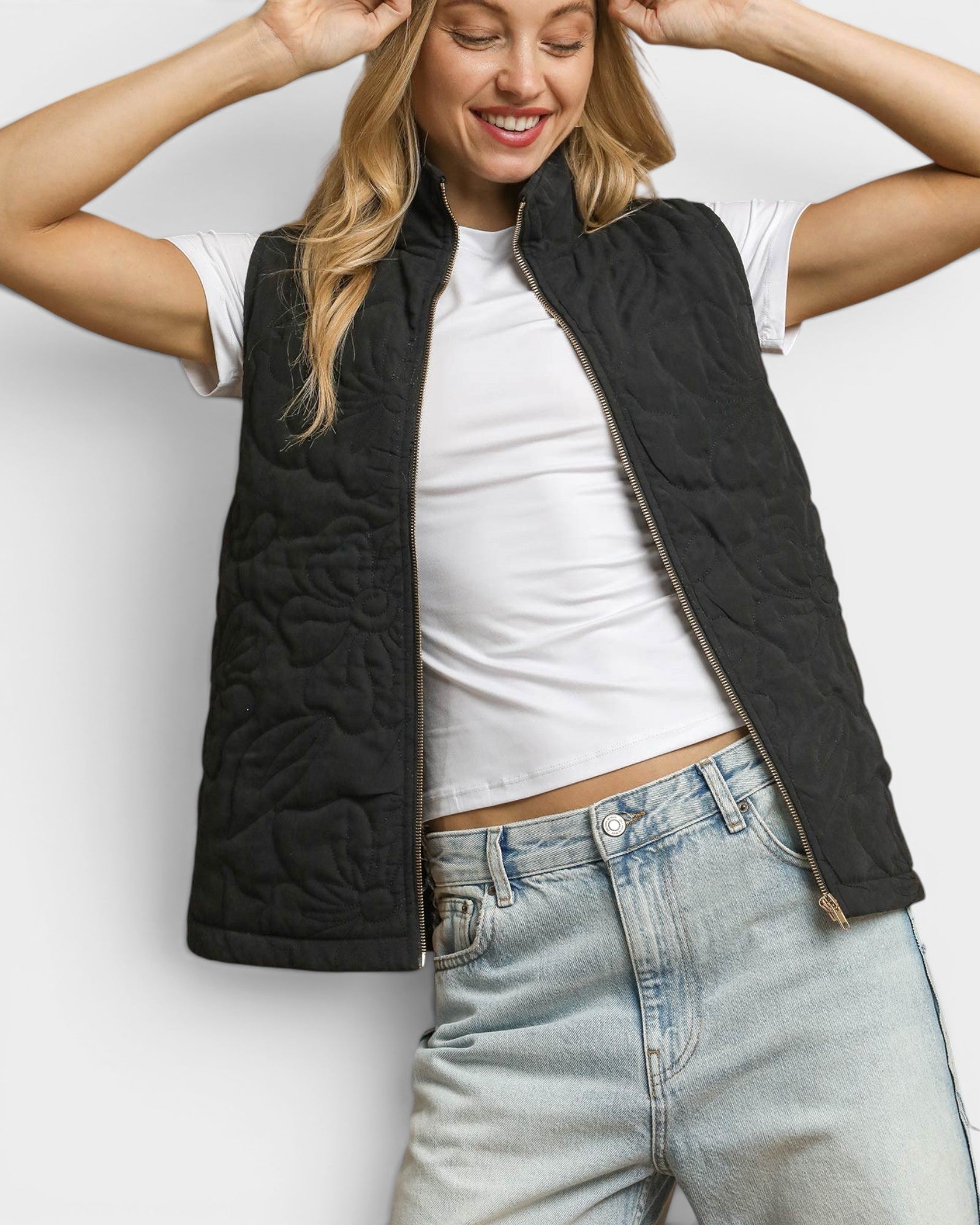 Woman wearing a black quilted vest over a white shirt and light blue jeans on a plain background