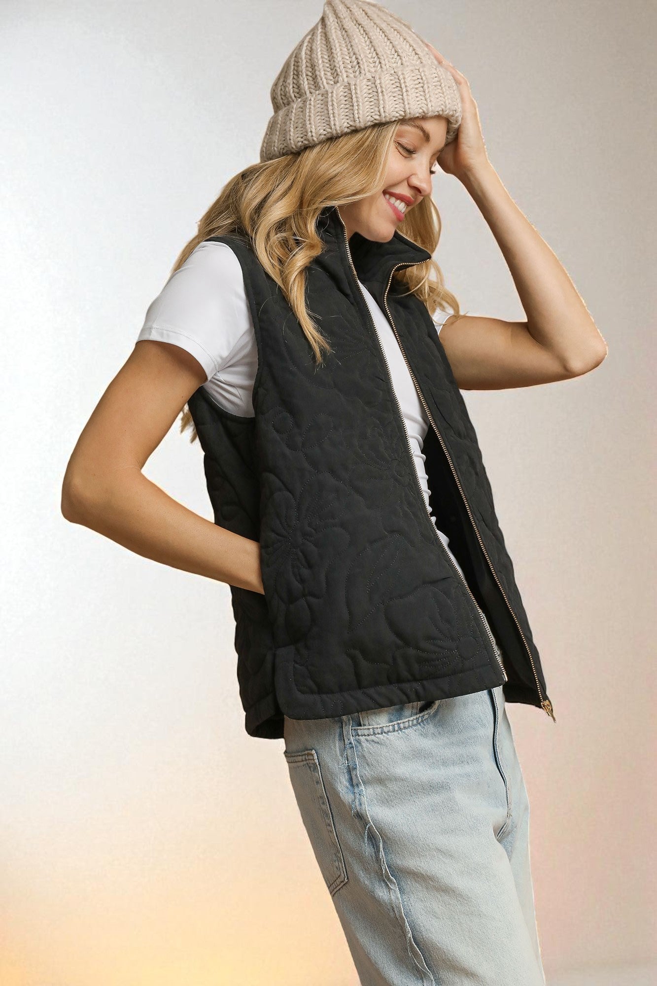 Woman wearing a black quilted vest, white shirt, light blue jeans, and beige knit beanie on a gradient background.