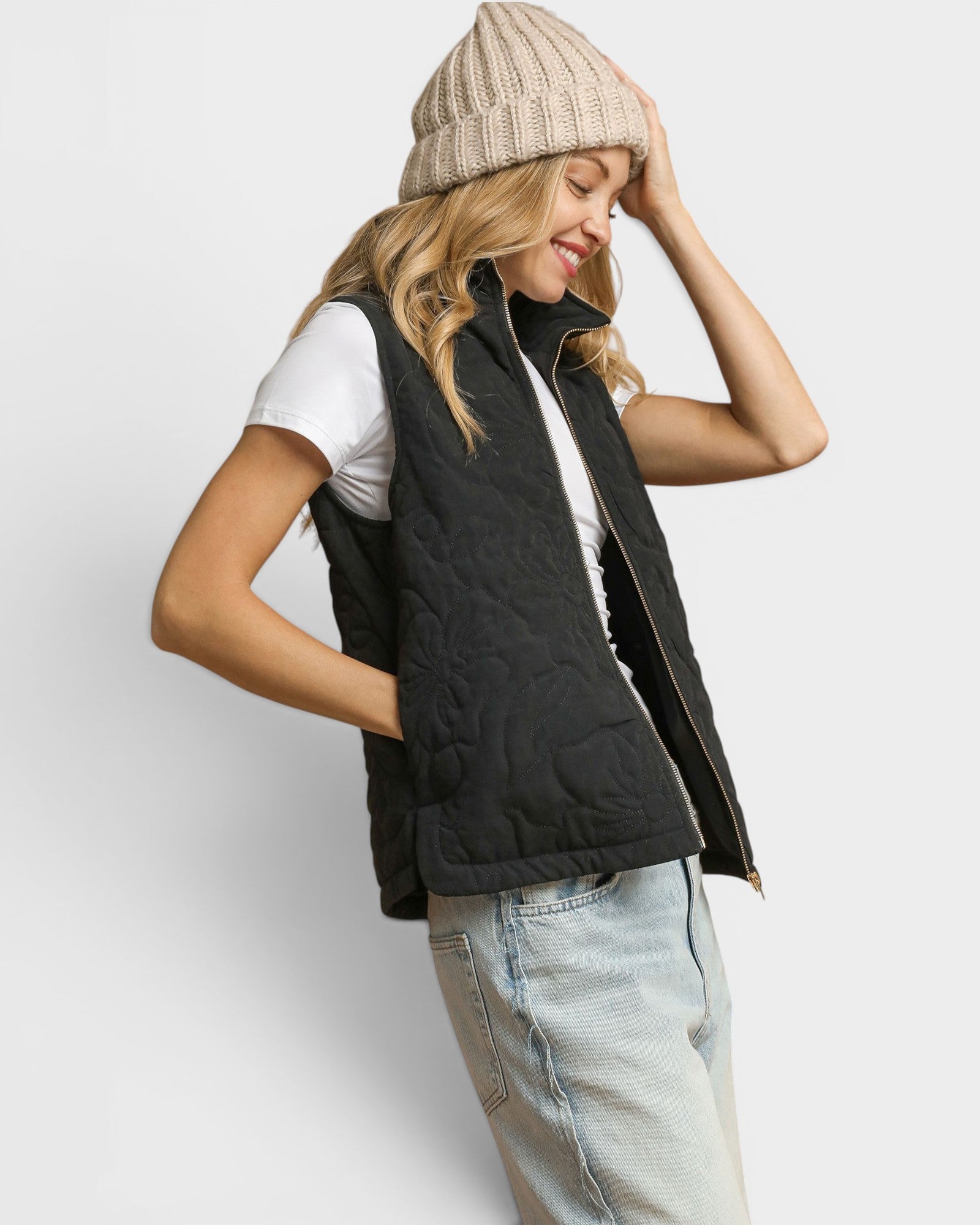 Woman wearing a black puffer vest, white shirt, light blue jeans, and beige knit beanie on a light gray background