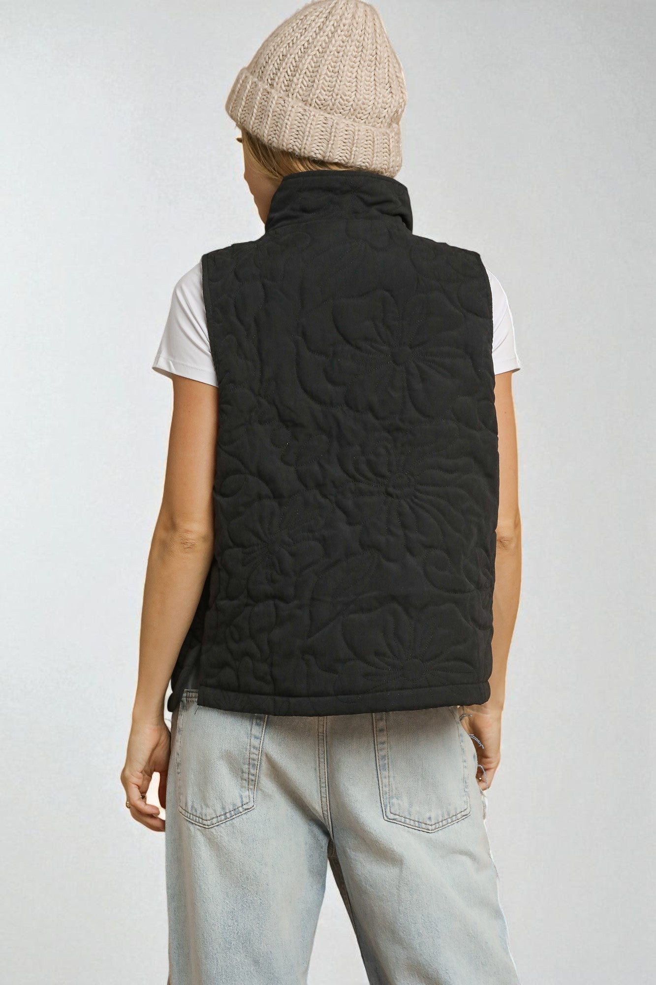 Person wearing a black quilted vest, white shirt, light blue jeans, and beige knit cap on a plain background