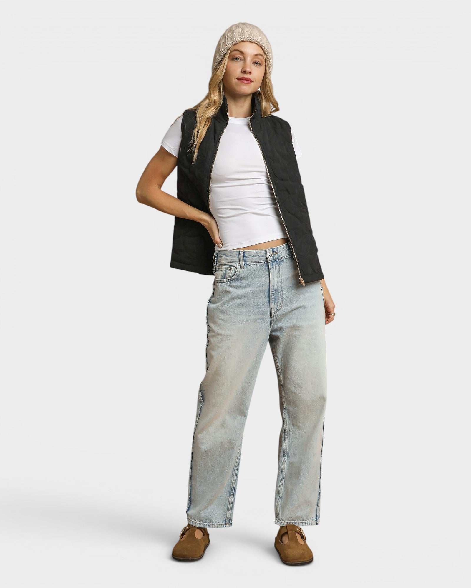Woman wearing a black vest, white shirt, light blue jeans, and brown shoes on a white background