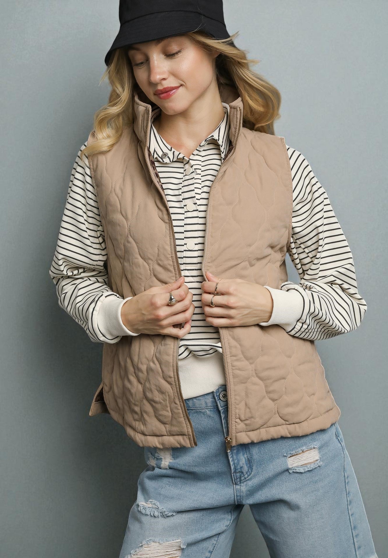 Woman wearing a beige quilted vest over a striped shirt with ripped jeans against a gray background