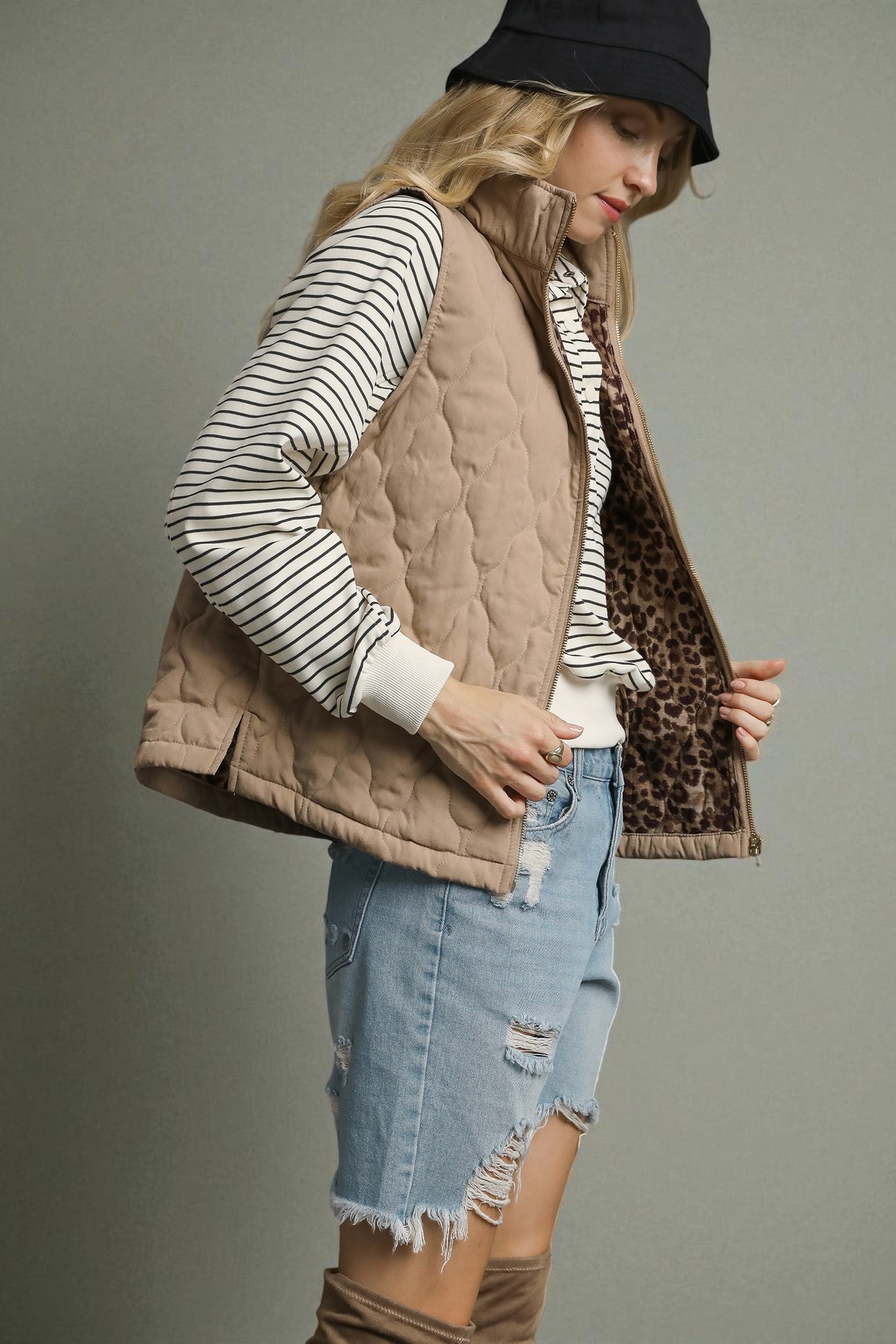 Person wearing a quilted vest with leopard print trim, striped shirt, denim shorts, and brown boots on a gray background.