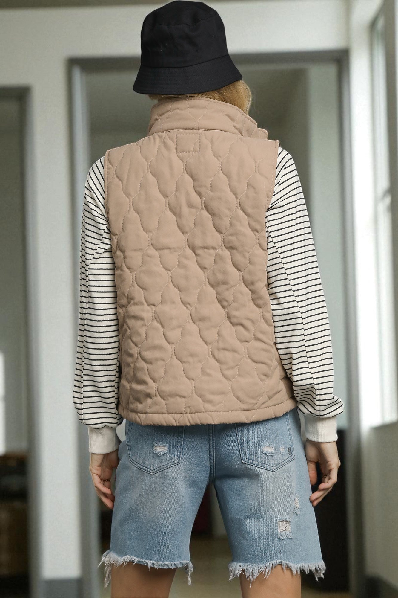 Person wearing a beige quilted vest over a striped shirt with denim shorts, standing indoors.