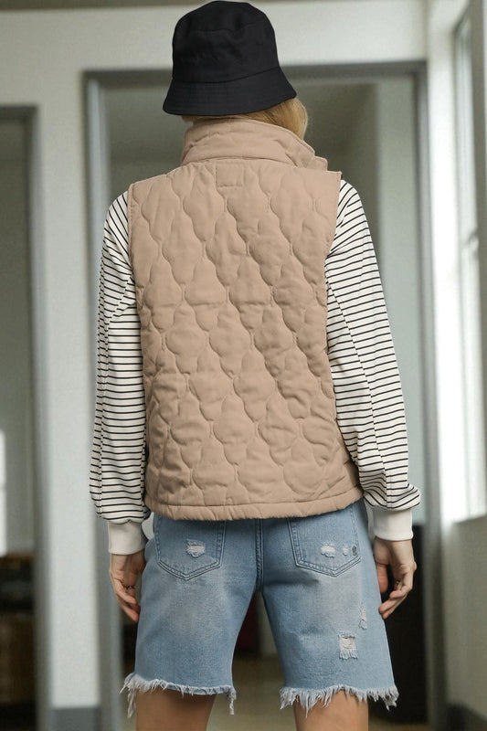 Person wearing a beige quilted vest over a striped shirt with denim shorts, standing indoors.