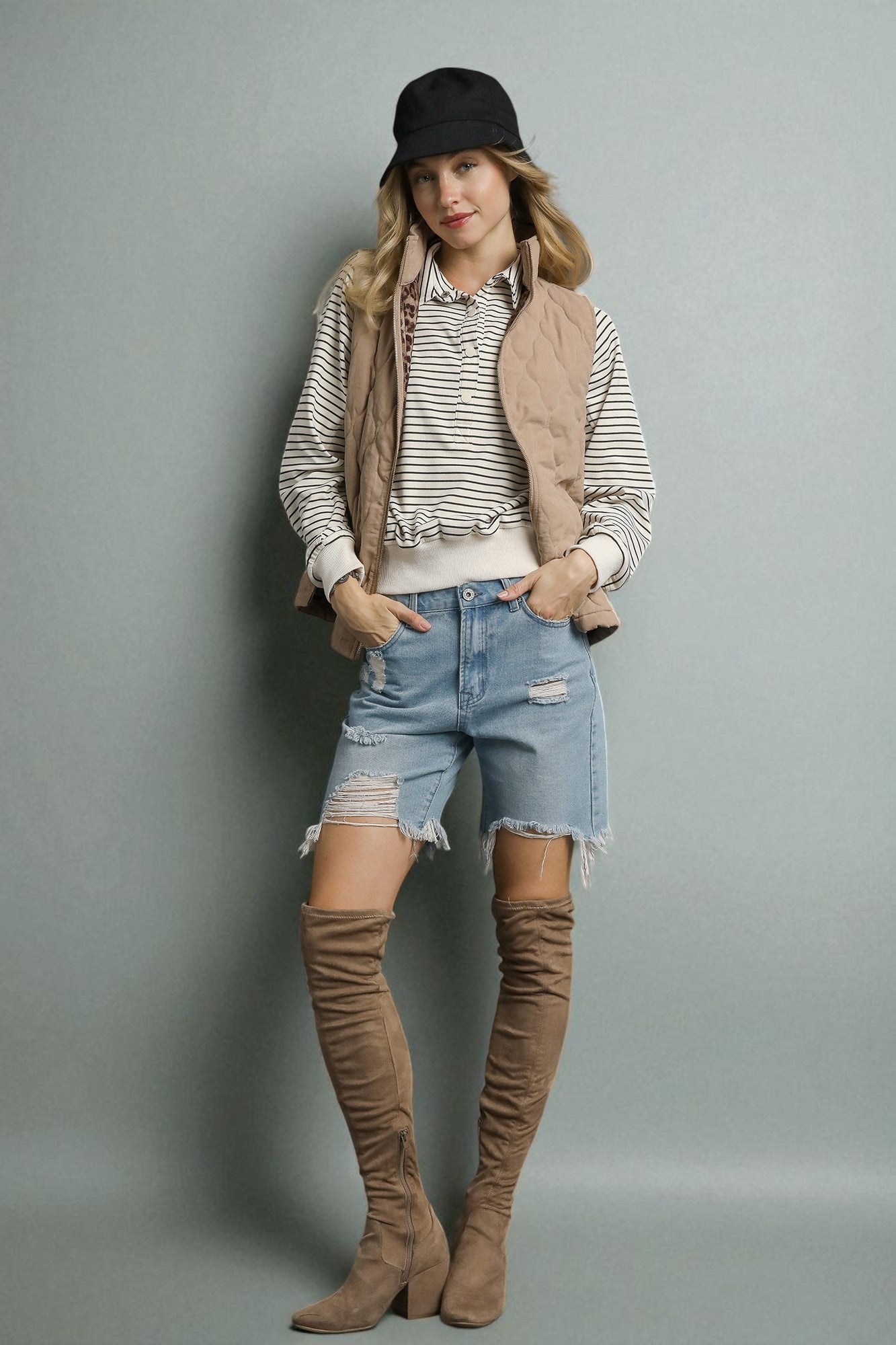 Woman wearing a striped shirt, denim shorts, brown suede boots, and a beige vest against a gray background.