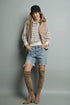 Woman wearing a striped shirt, denim shorts, brown suede boots, and a beige vest against a gray background.