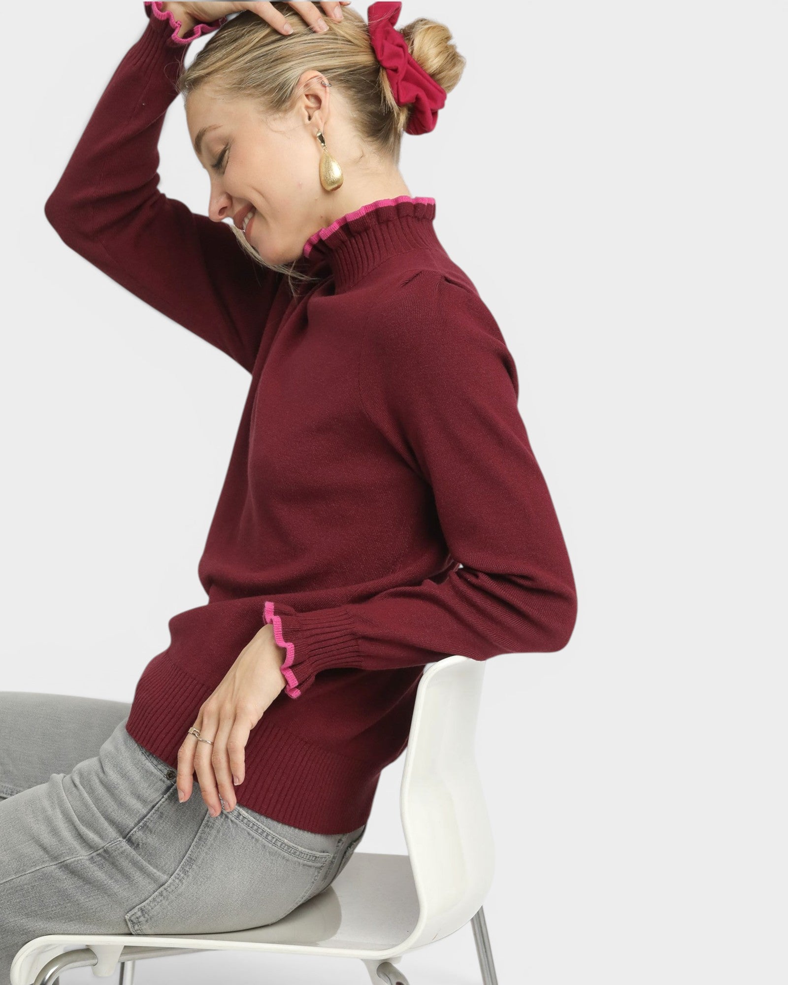 Woman wearing a burgundy sweater with pink trim sitting on a white chair against a light gray background