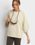 Woman wearing a beige sweater holding a black necklace with a red pendant on a light gray background