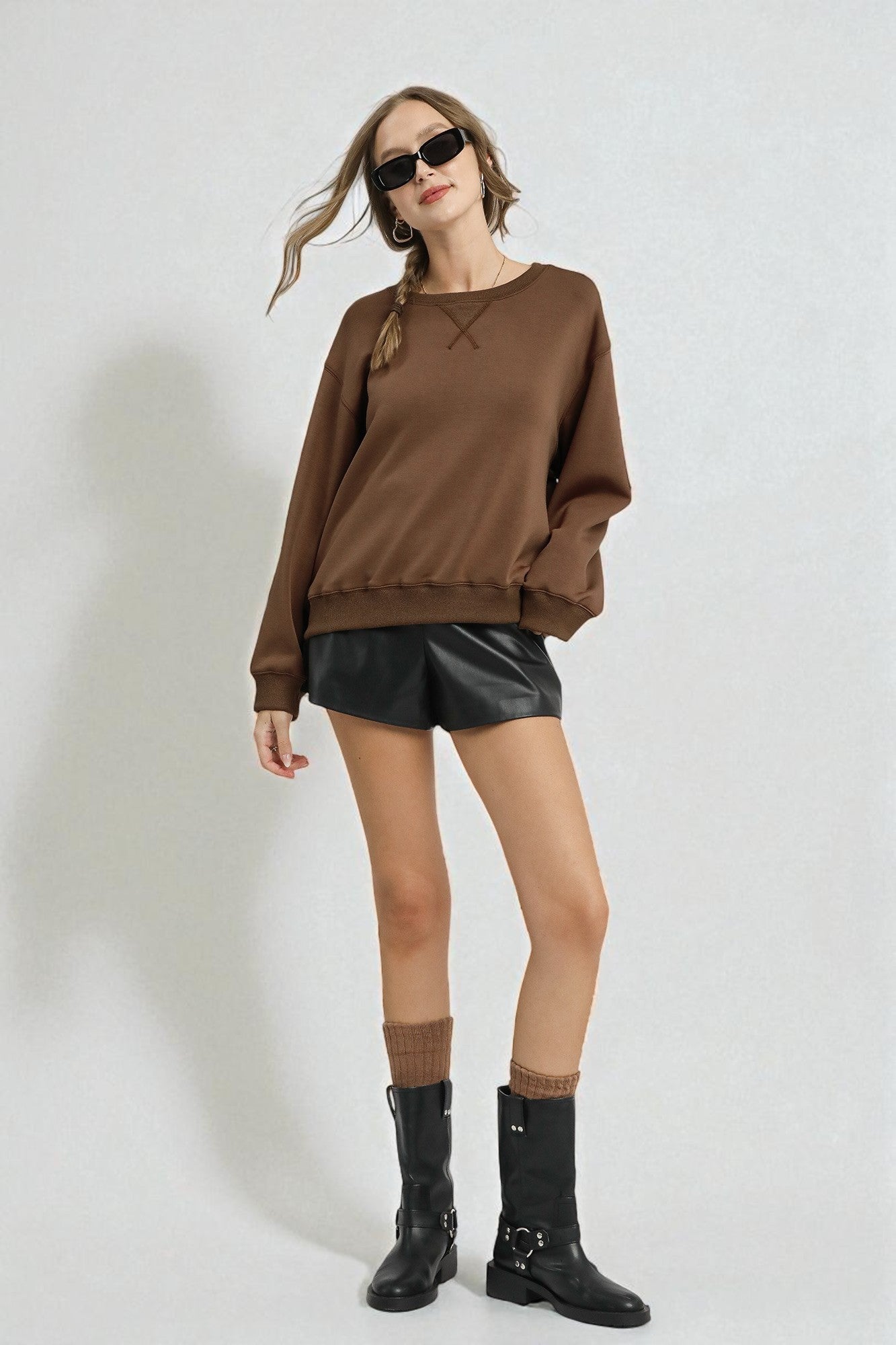 Woman wearing a brown sweater, black leather shorts, and black boots on a white background