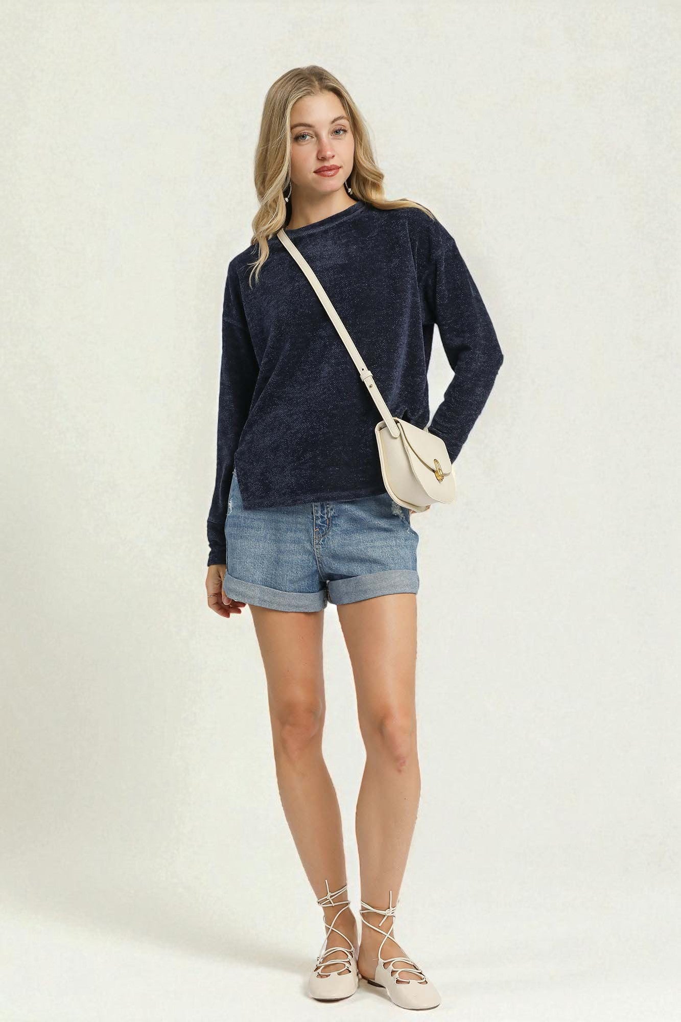Woman wearing a navy sweater and denim shorts with a white bag on a plain background
