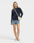 Woman wearing a navy sweater and denim shorts with a white bag on a light gray background