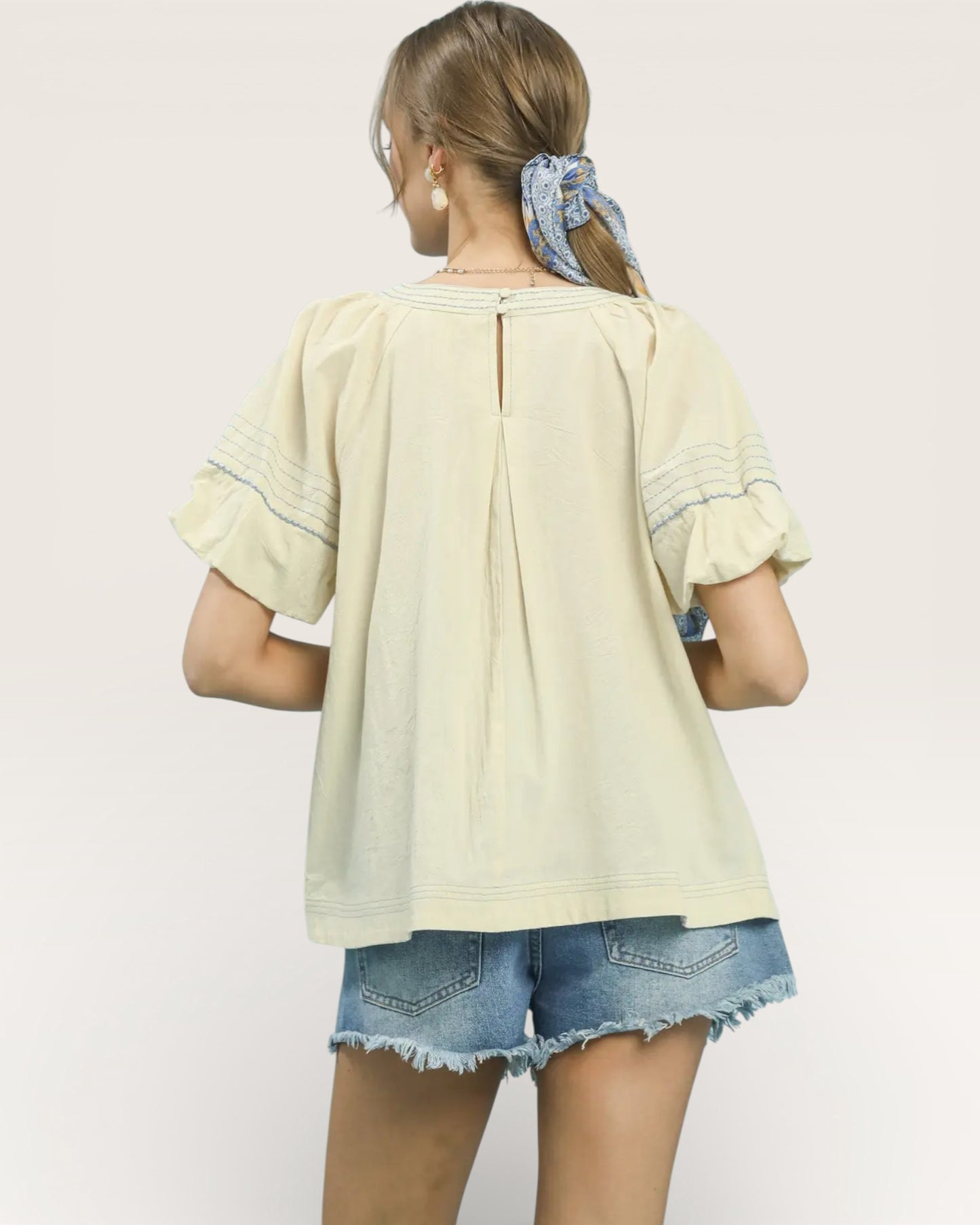 Woman wearing a light-colored blouse with ruffled sleeves and denim shorts on a plain background