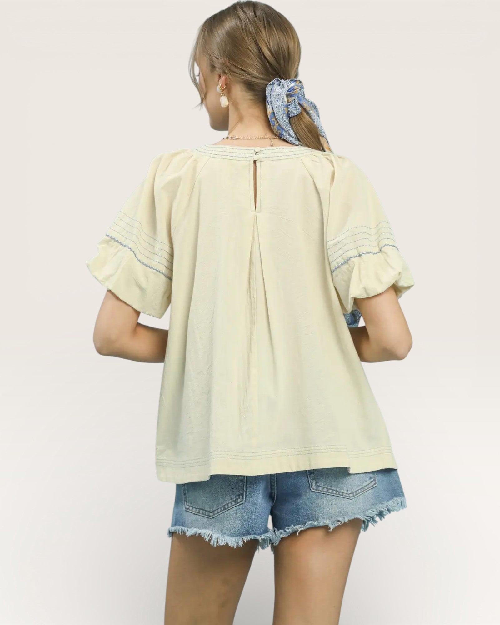 Woman wearing a light-colored blouse with ruffled sleeves and denim shorts on a plain background