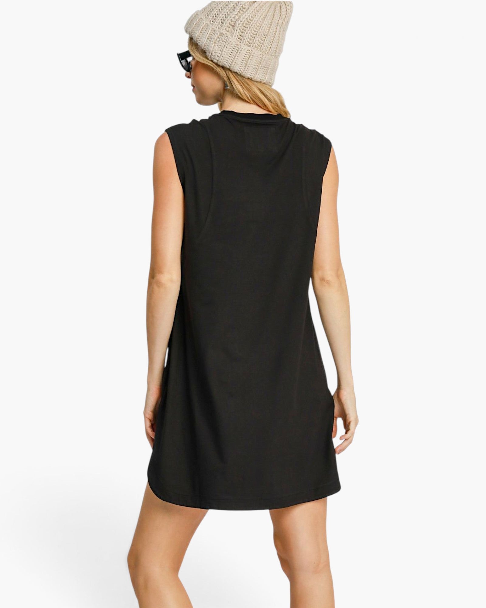 A woman shown from the back wearing a sleeveless black tunic dress, a beige knit beanie, and black sunglasses against a plain white studio background.