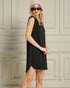 Woman wearing a black sleeveless dress and sunglasses against a beige paneled wall.
