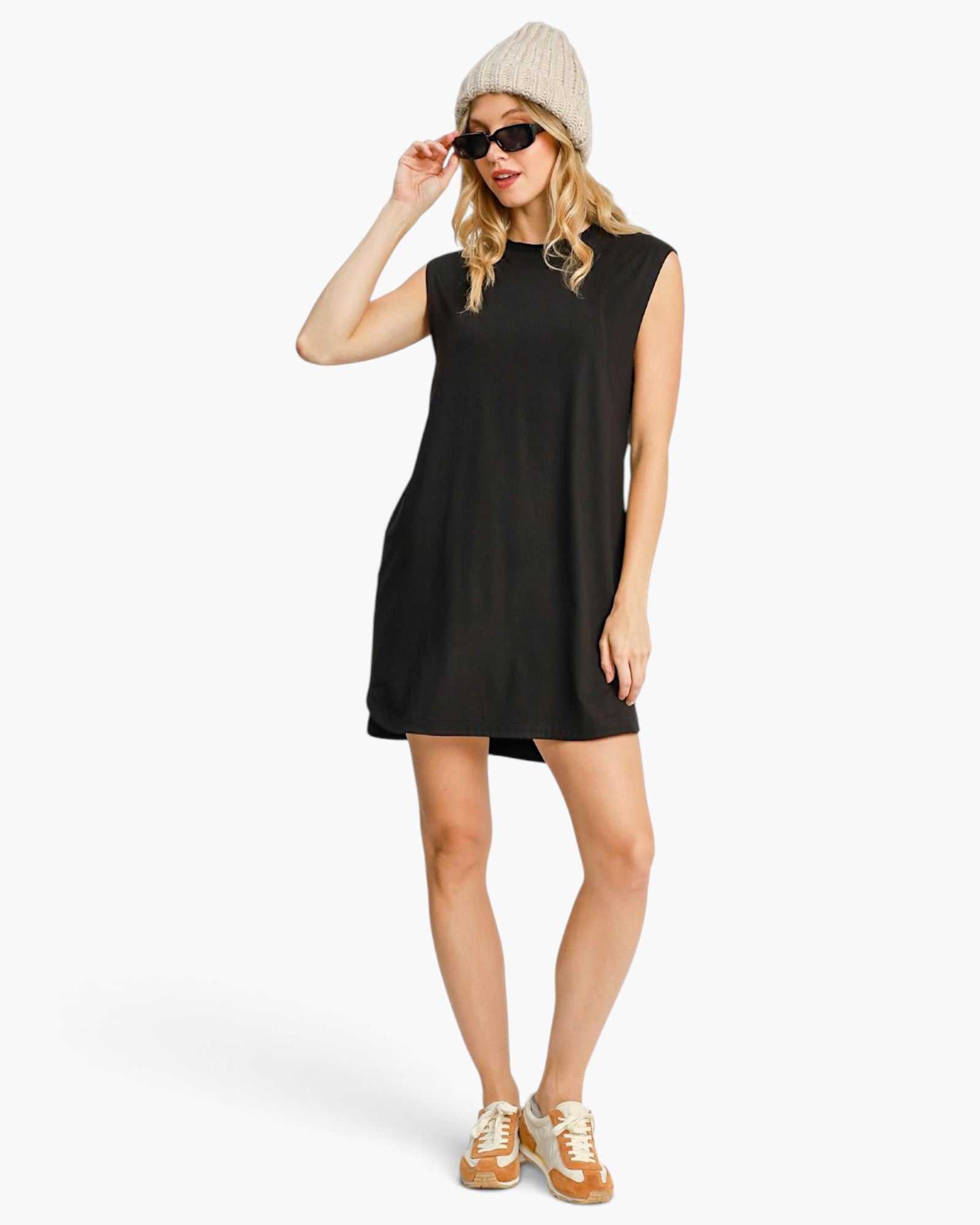 A woman modeling a sleeveless black shift dress, styled with a beige knit beanie, black sunglasses, and tan sneakers, against a plain white studio background.