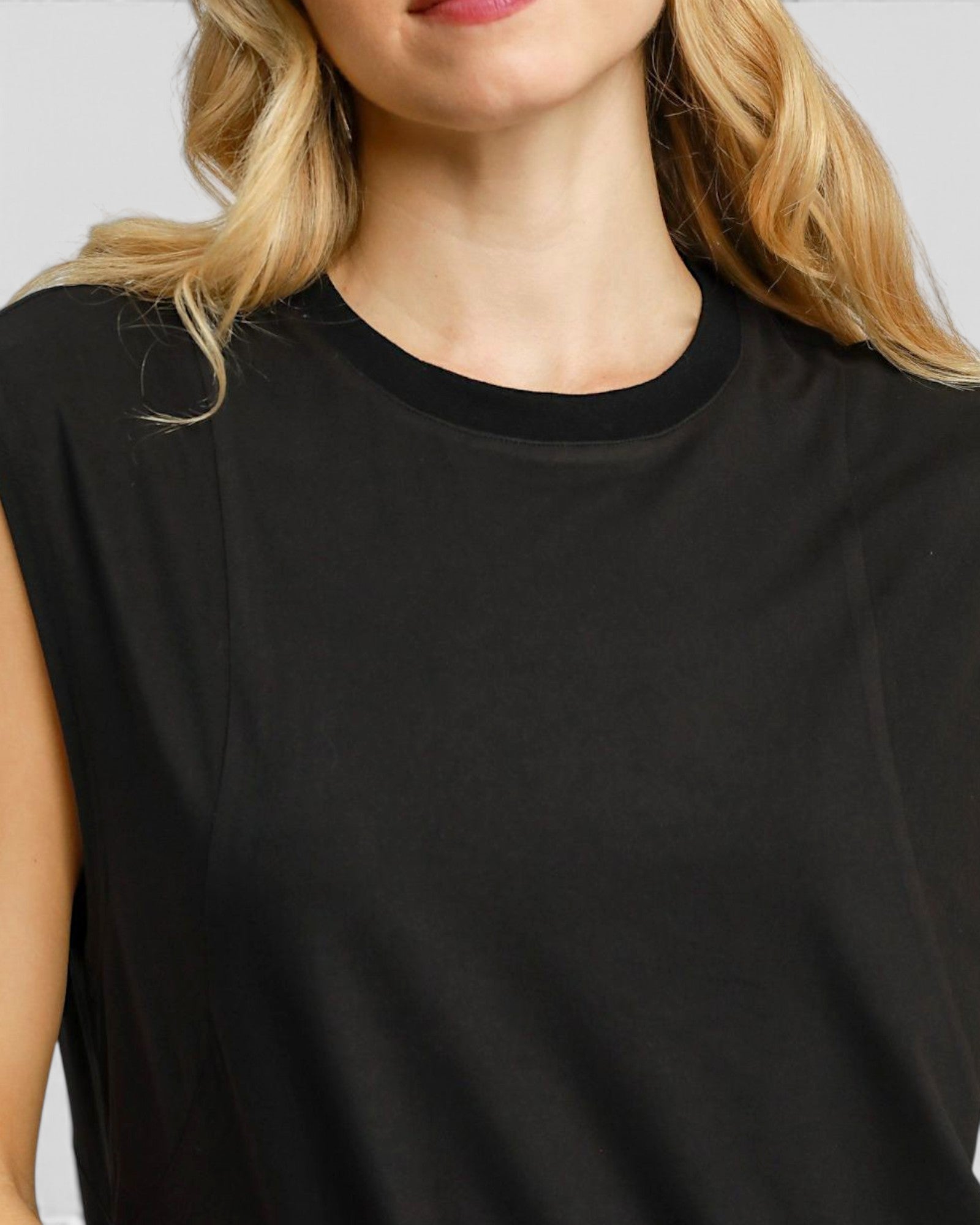 Close-up of a woman wearing a black sleeveless top with a ribbed crew neck, showing her blonde hair and shoulder against a light gray background.