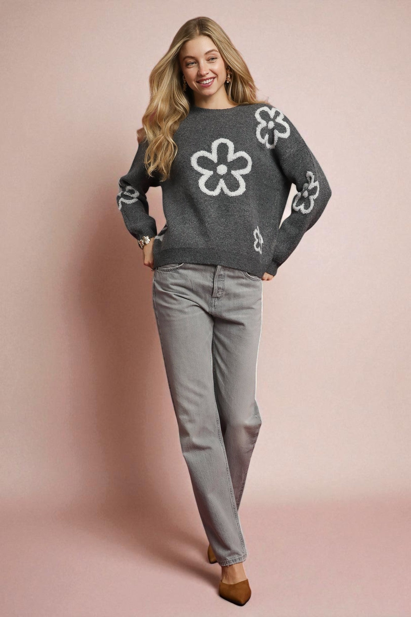 Woman wearing a dark gray sweater with floral patterns and light gray pants against a pink background