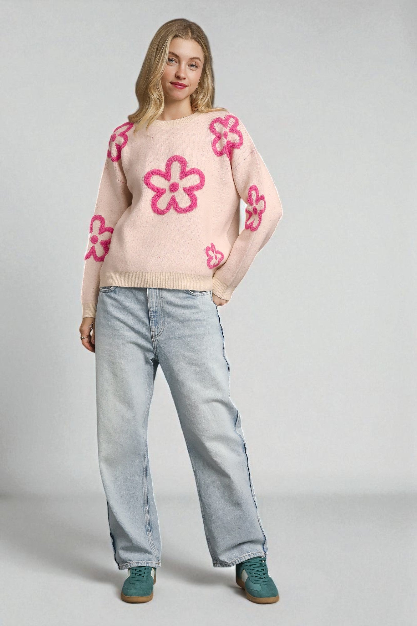 Woman wearing a light pink sweater with pink floral patterns and light blue jeans on a gray background