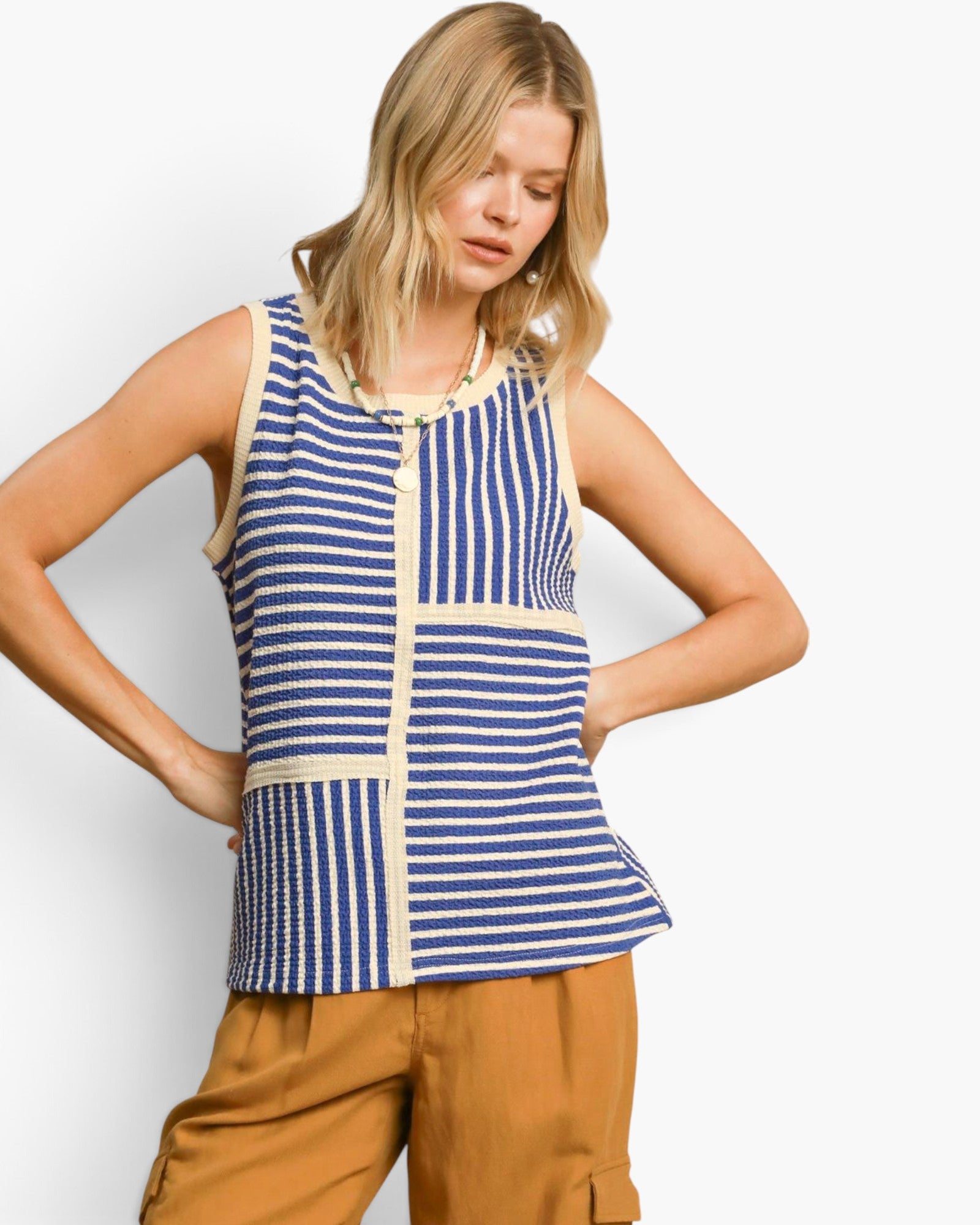 A woman modeling a sleeveless knit top with blue and cream vertical stripes and patchwork panel details, paired with tan cargo pants, against a light gray studio background.