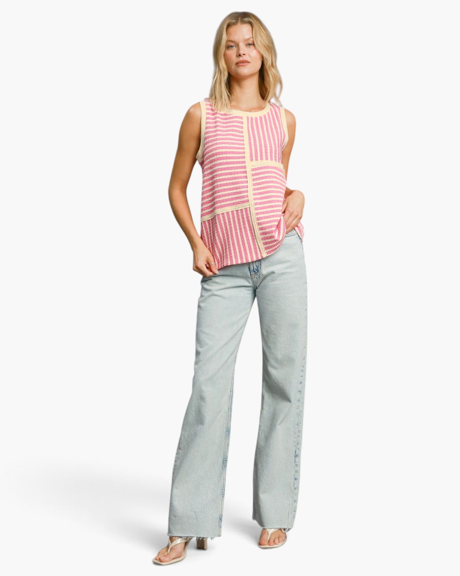 A woman modeling a sleeveless knit top with pink and white vertical stripes and cream trim, paired with light-wash wide-leg jeans, against a plain white studio background.