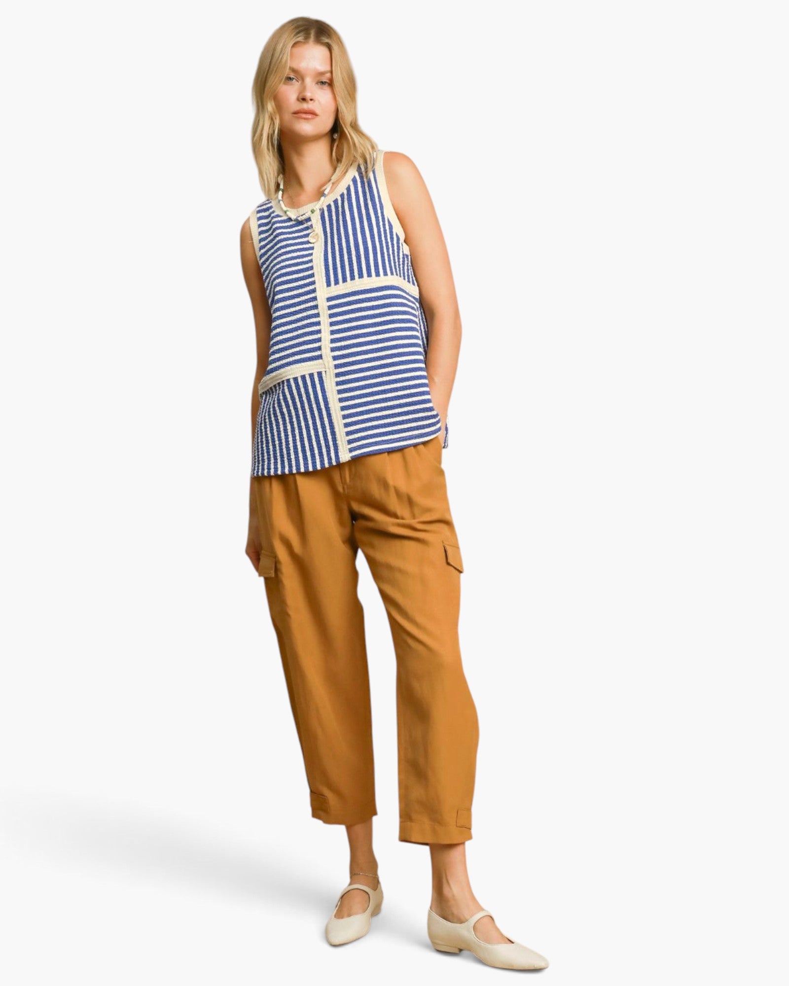 A woman modeling a sleeveless top with blue and white stripes and cream trim, paired with mustard-brown cropped pants and cream flats, against a light gray studio background.
