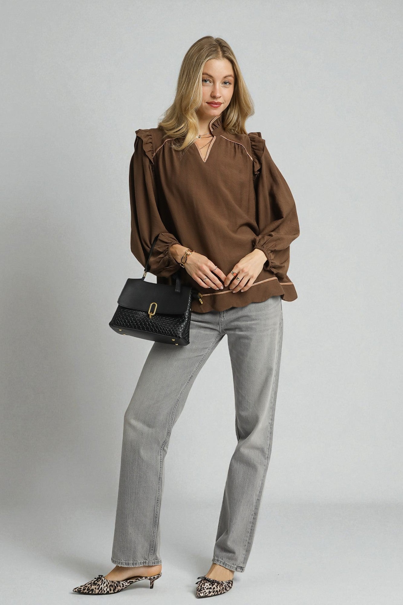 Woman wearing a brown blouse and gray pants holding a black handbag on a plain background