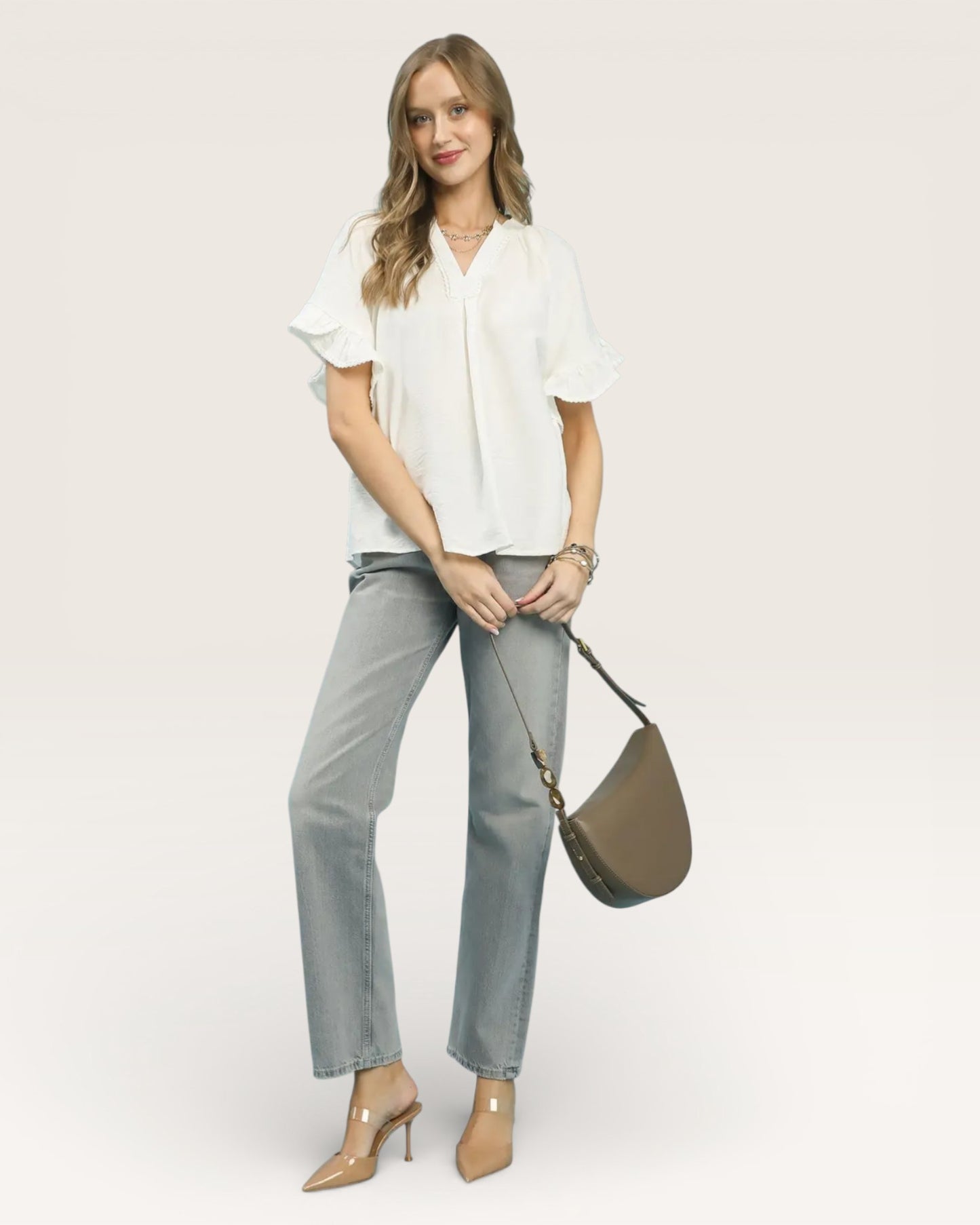 Woman wearing a white blouse and gray pants, holding a brown handbag on a plain background