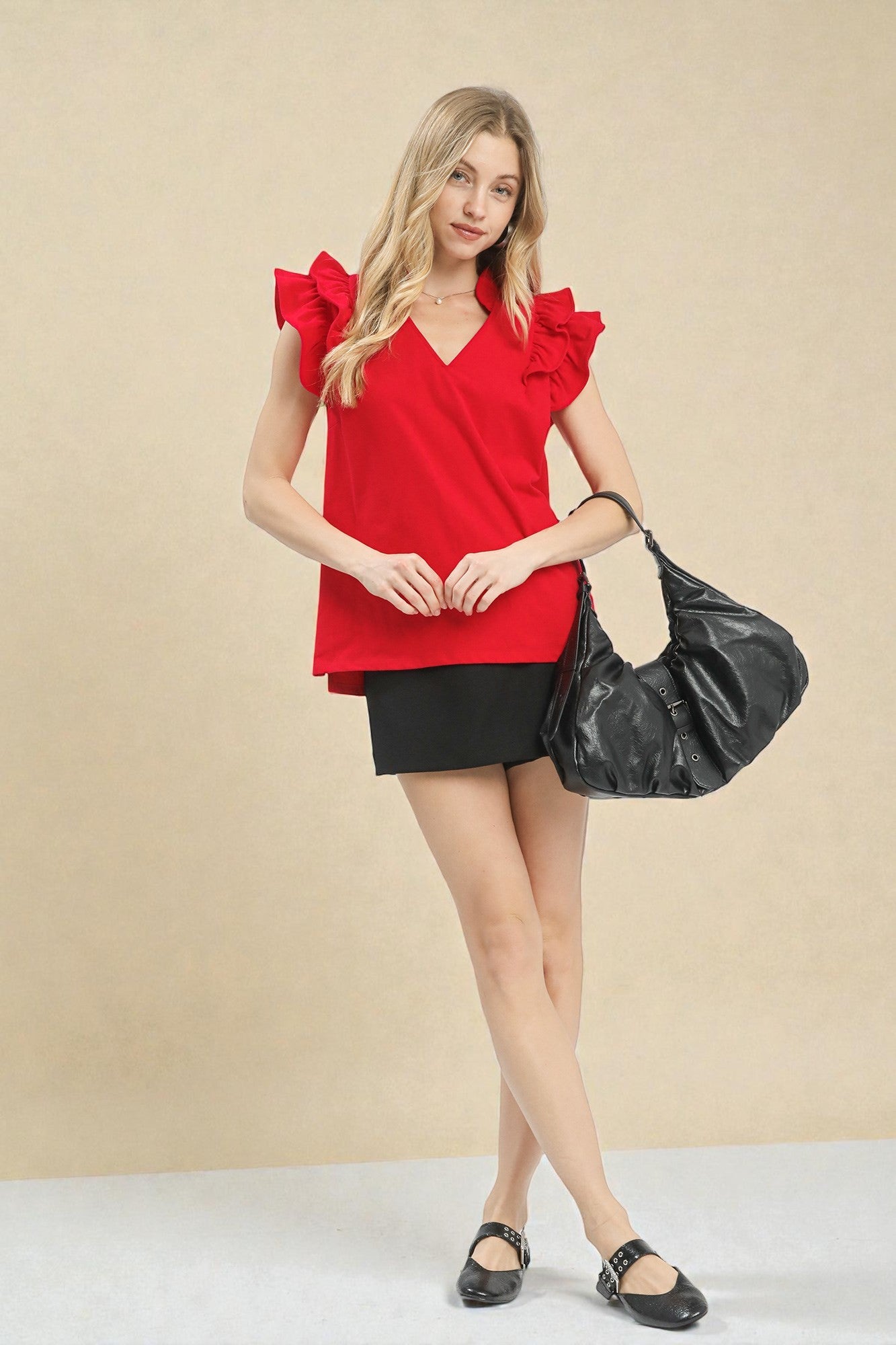 Woman wearing a red blouse with ruffled sleeves and a black skirt, holding a black handbag against a beige background.