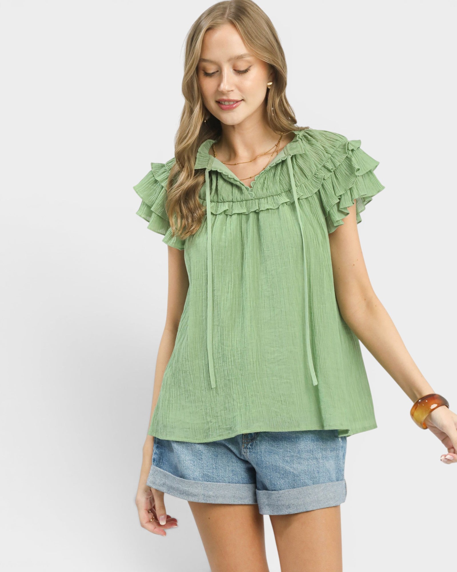 Woman wearing a green blouse with ruffled sleeves and denim shorts on a white background