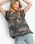 A woman wearing a black short-sleeve blouse with a beige toile-style landscape print and ruffled trim, paired with light blue jeans, posing against a white studio background.