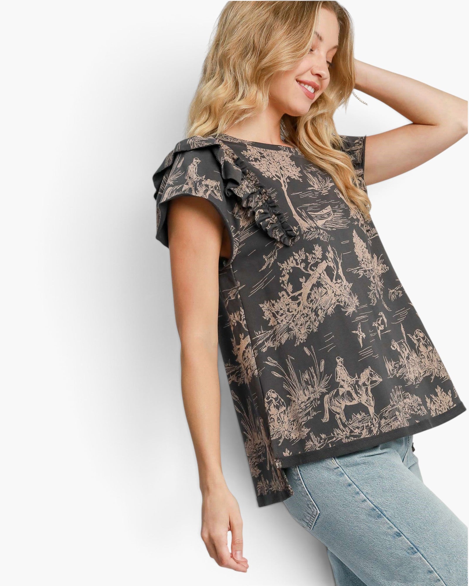 A woman wearing a black short-sleeve blouse with beige toile de Jouy print and ruffled sleeves, paired with light blue jeans, posing against a white studio background.