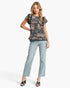 A woman modeling a short-sleeve black top with a beige toile-style landscape print, paired with light-wash blue jeans and black sandals, against a plain white studio background.