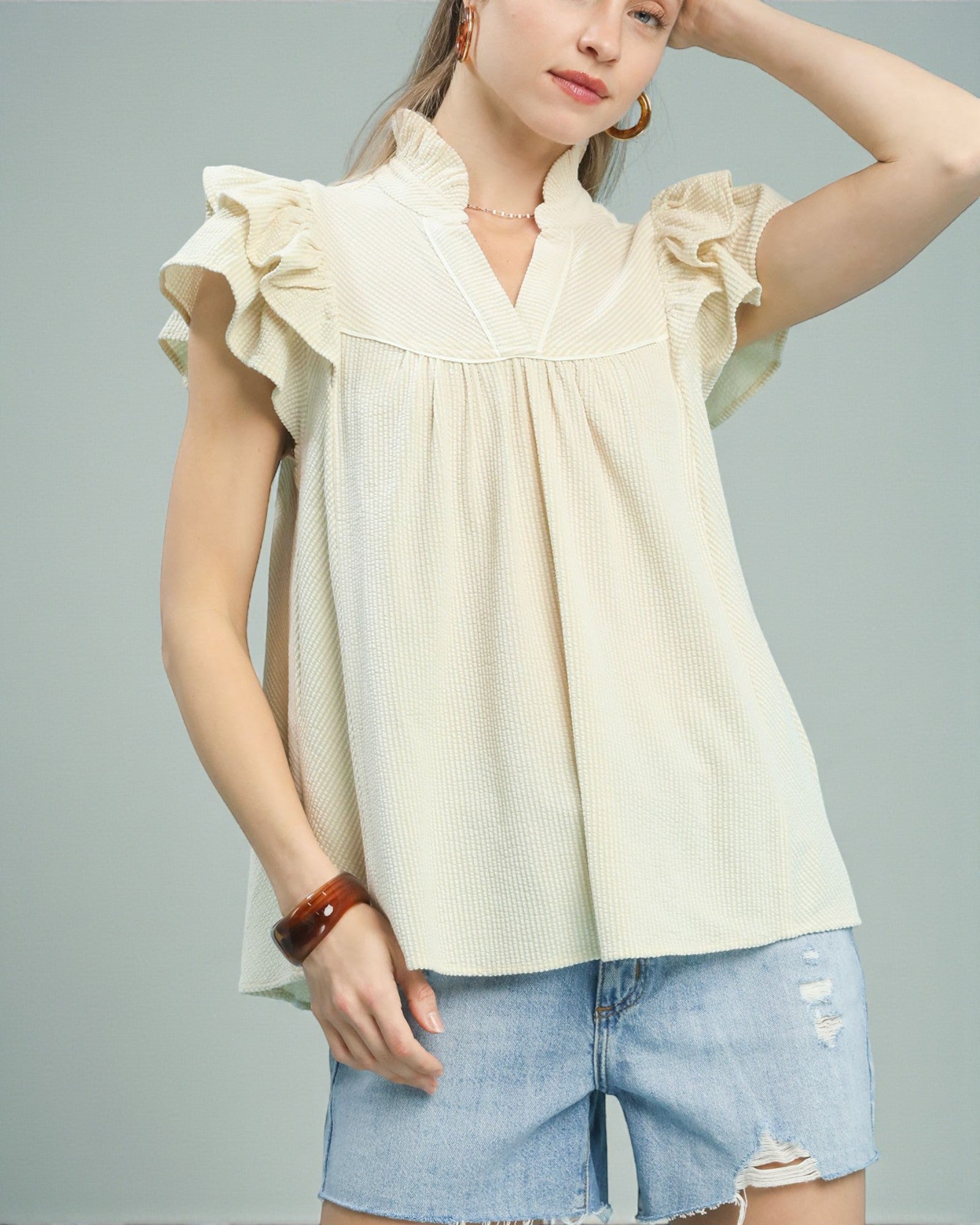 Woman wearing a light yellow blouse with ruffled sleeves and denim shorts on a gray background