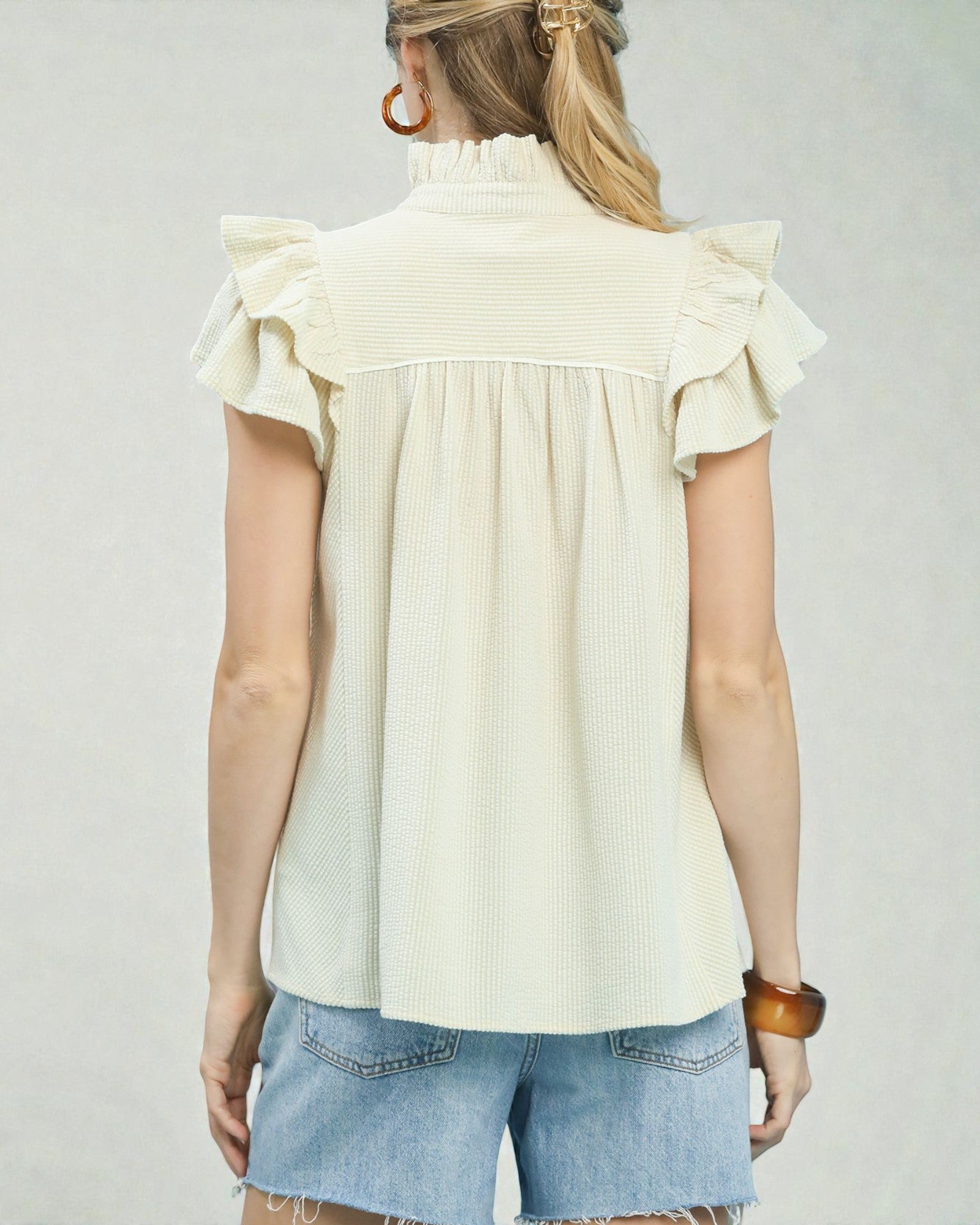 Woman wearing a cream blouse with ruffled shoulders and denim shorts on a plain background