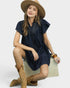 Woman wearing a navy dress, beige hat, and tan boots sitting on a white box against a light gray background.