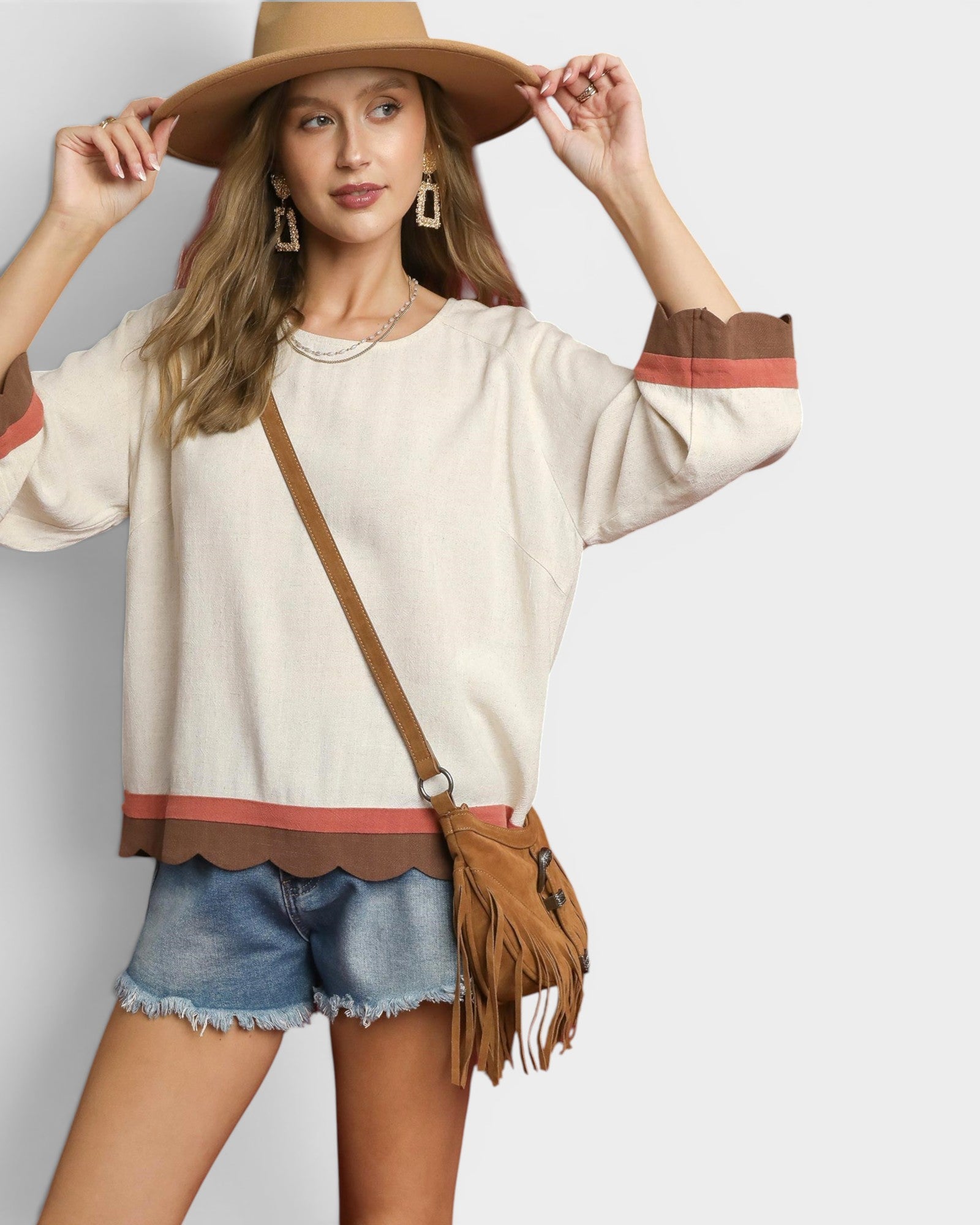 Woman wearing a beige sweater with brown and red accents, denim shorts, and a tan hat on a light gray background.