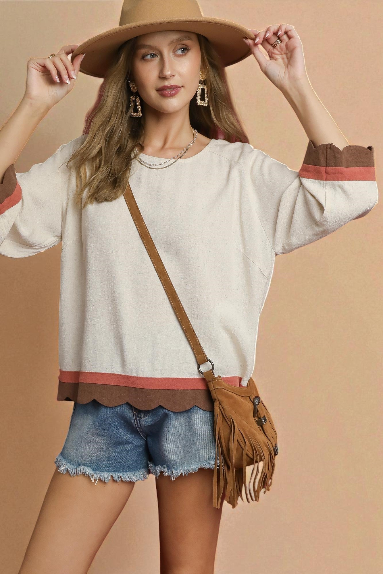 Woman wearing a beige top with scalloped hem, denim shorts, and a wide-brimmed hat, holding a brown leather bag.