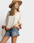 Woman wearing a beige top with brown accents and denim shorts on a white background
