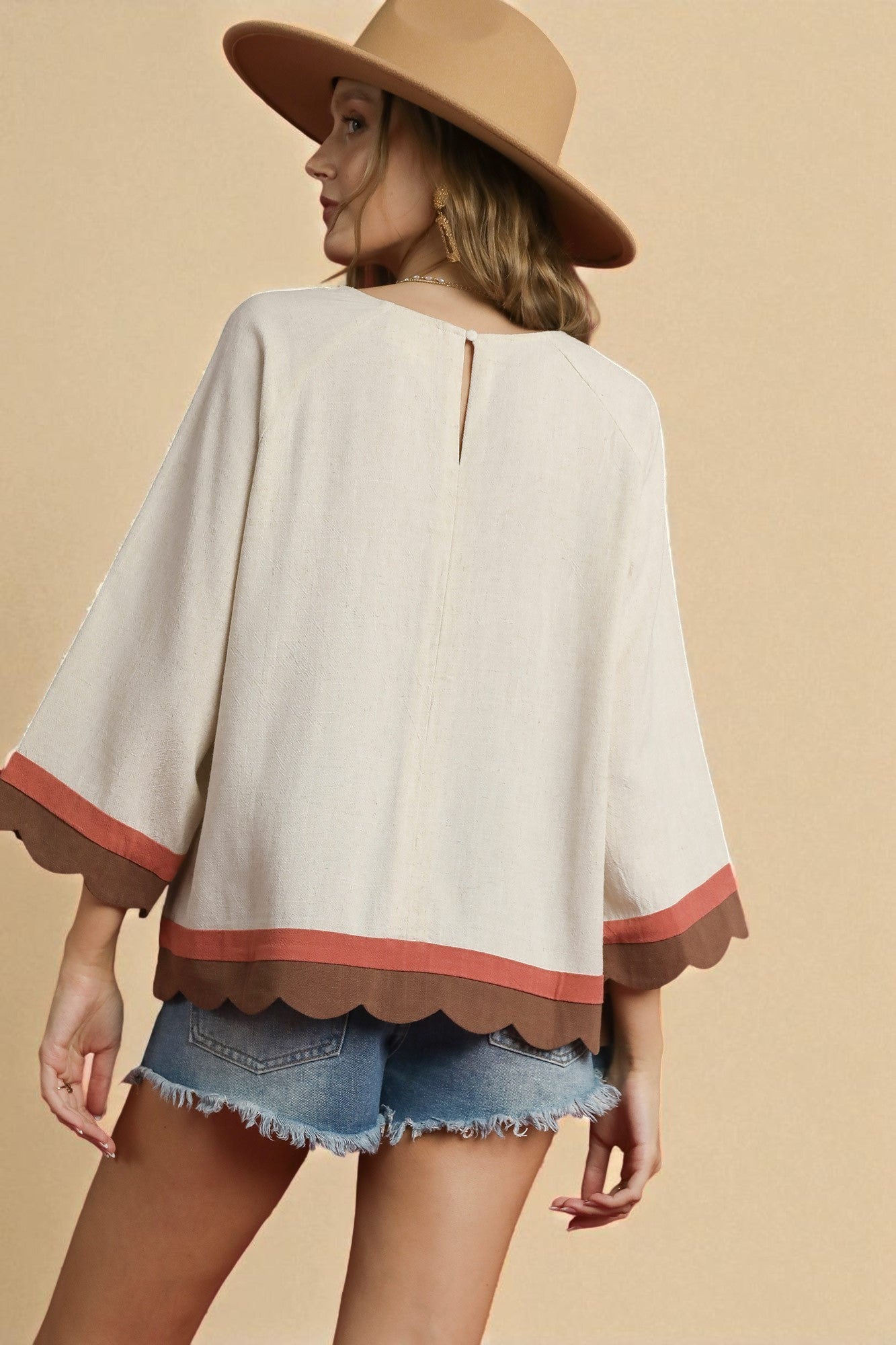 Woman wearing a beige blouse with red trim, denim shorts, and a wide-brimmed hat on a beige background