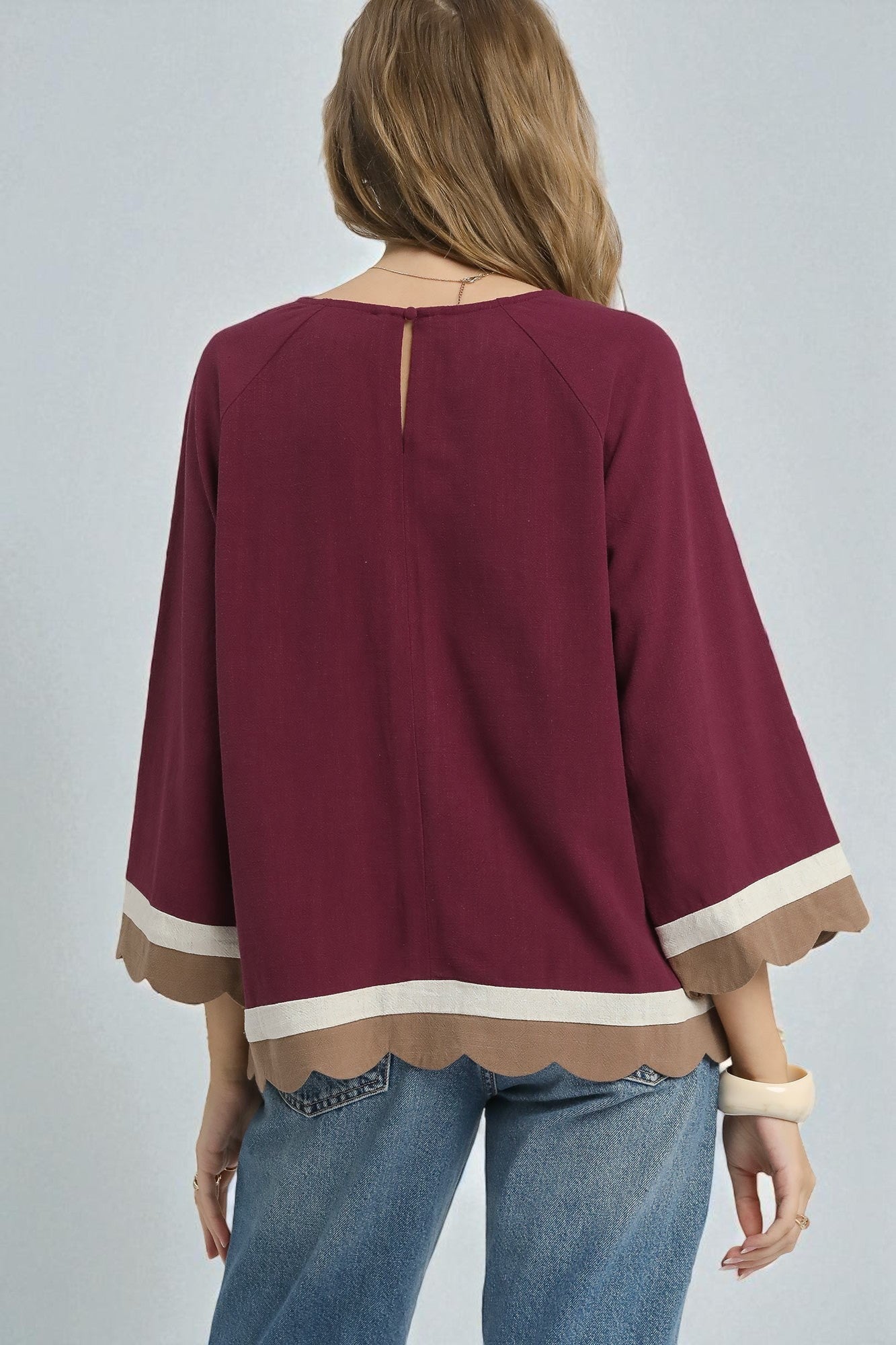 Maroon blouse with scalloped hem and cuffs worn by a person on a gray background