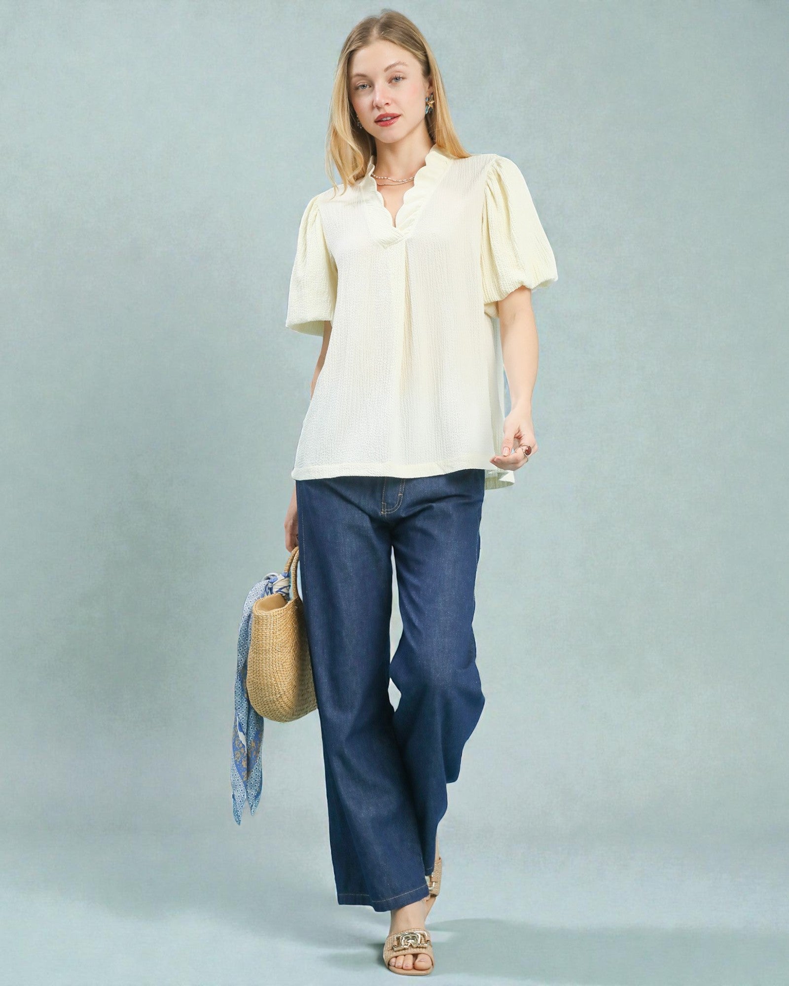 Woman wearing a light yellow blouse and blue pants, holding a woven bag on a plain background