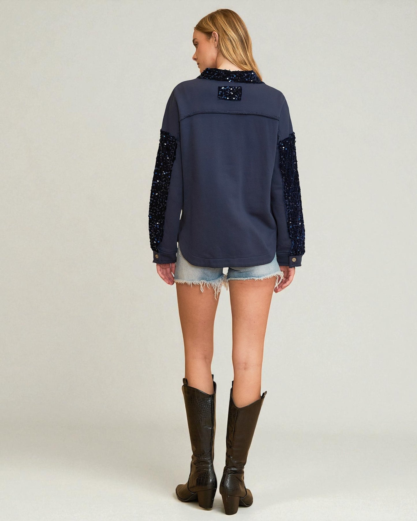 Woman wearing a navy blue blouse with sequin details on sleeves and back, denim shorts, and brown boots against a white background.