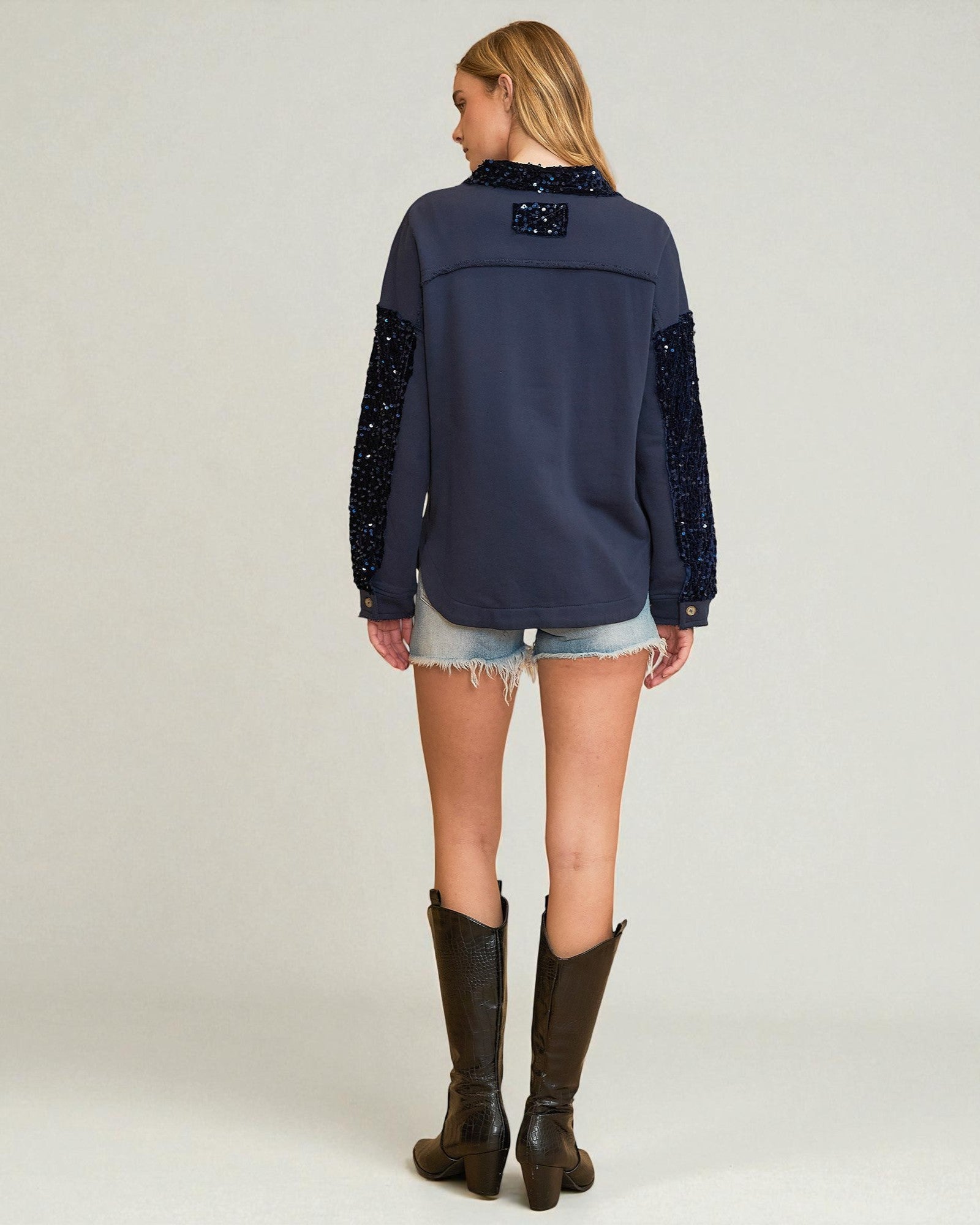 Woman wearing a navy blue blouse with sequin details on sleeves and back, denim shorts, and brown boots against a white background.