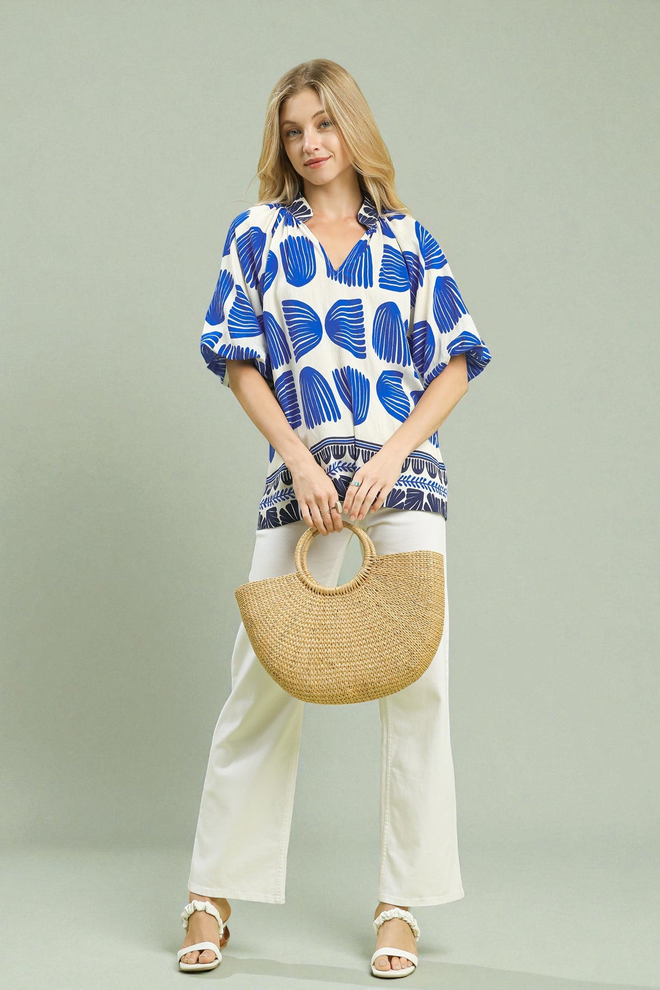 Woman wearing a blue and white patterned shirt with white pants, holding a straw bag on a light gray background