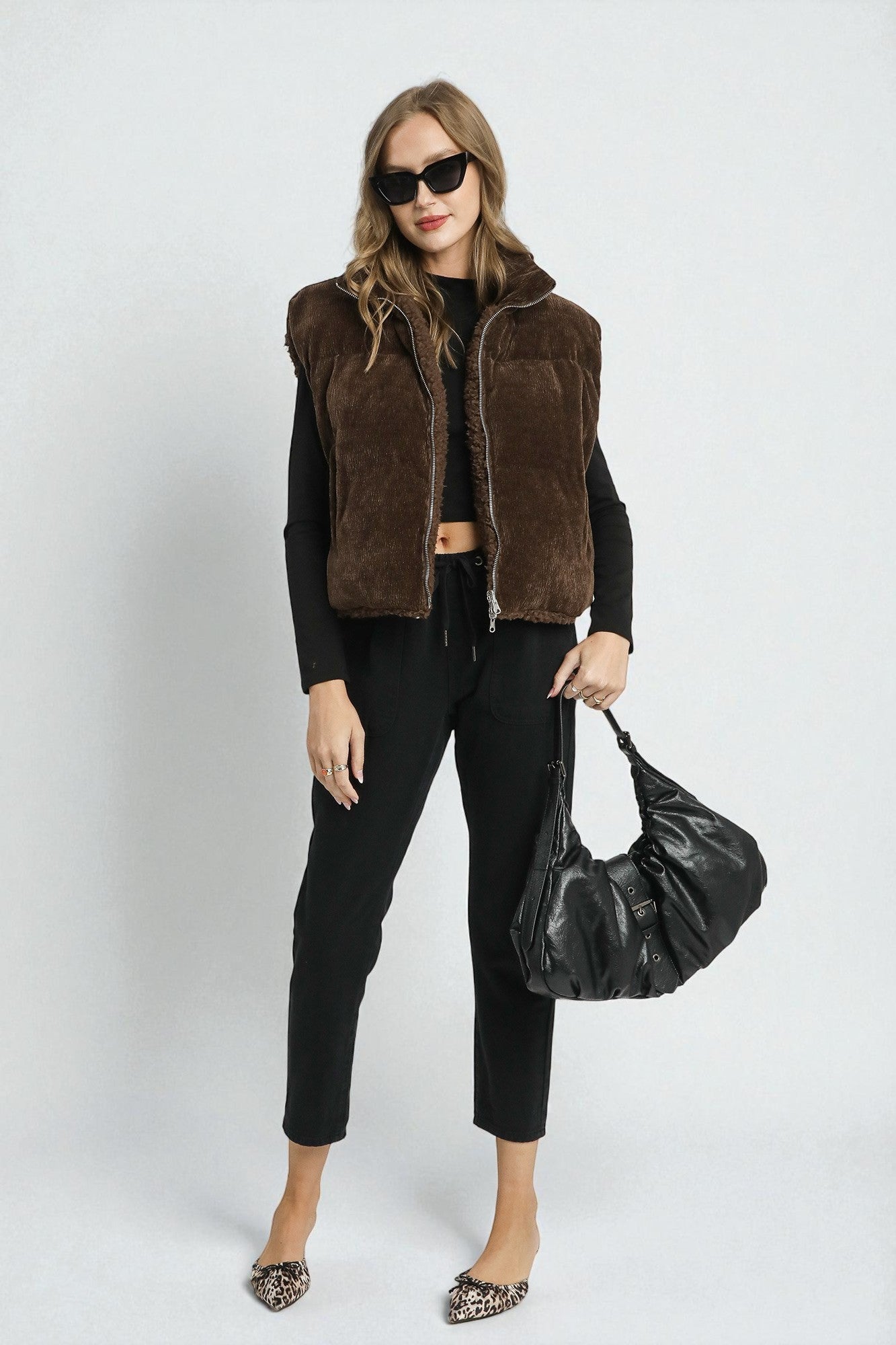 Woman wearing a brown vest, black top, and pants, holding a black handbag on a white background