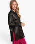 A woman modeling a black faux-leather button-up shirt with a satin-like sheen, paired with bright pink pleated pants, against a plain white studio background.