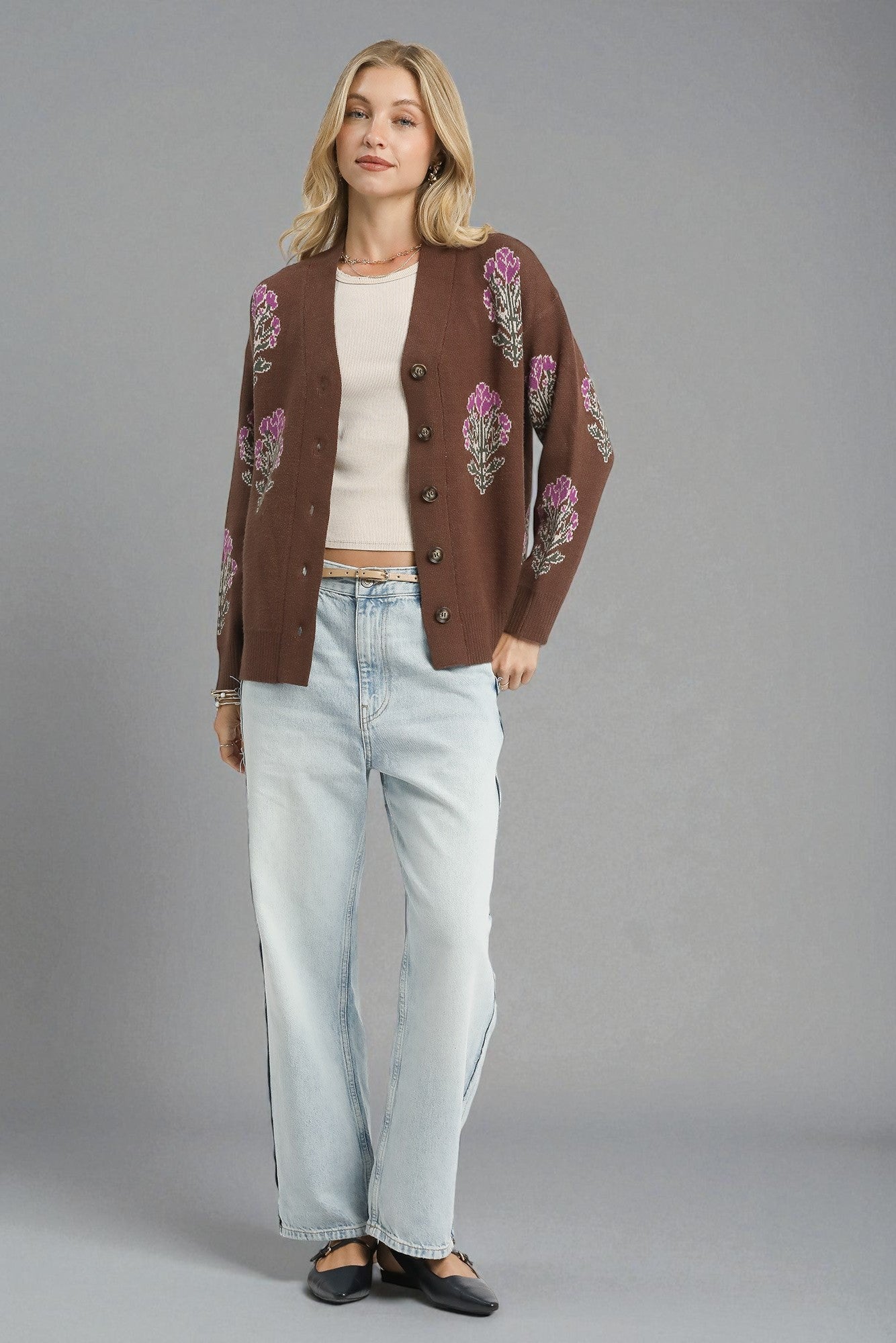 Woman wearing a brown embroidered cardigan, white top, and light blue jeans on a gray background