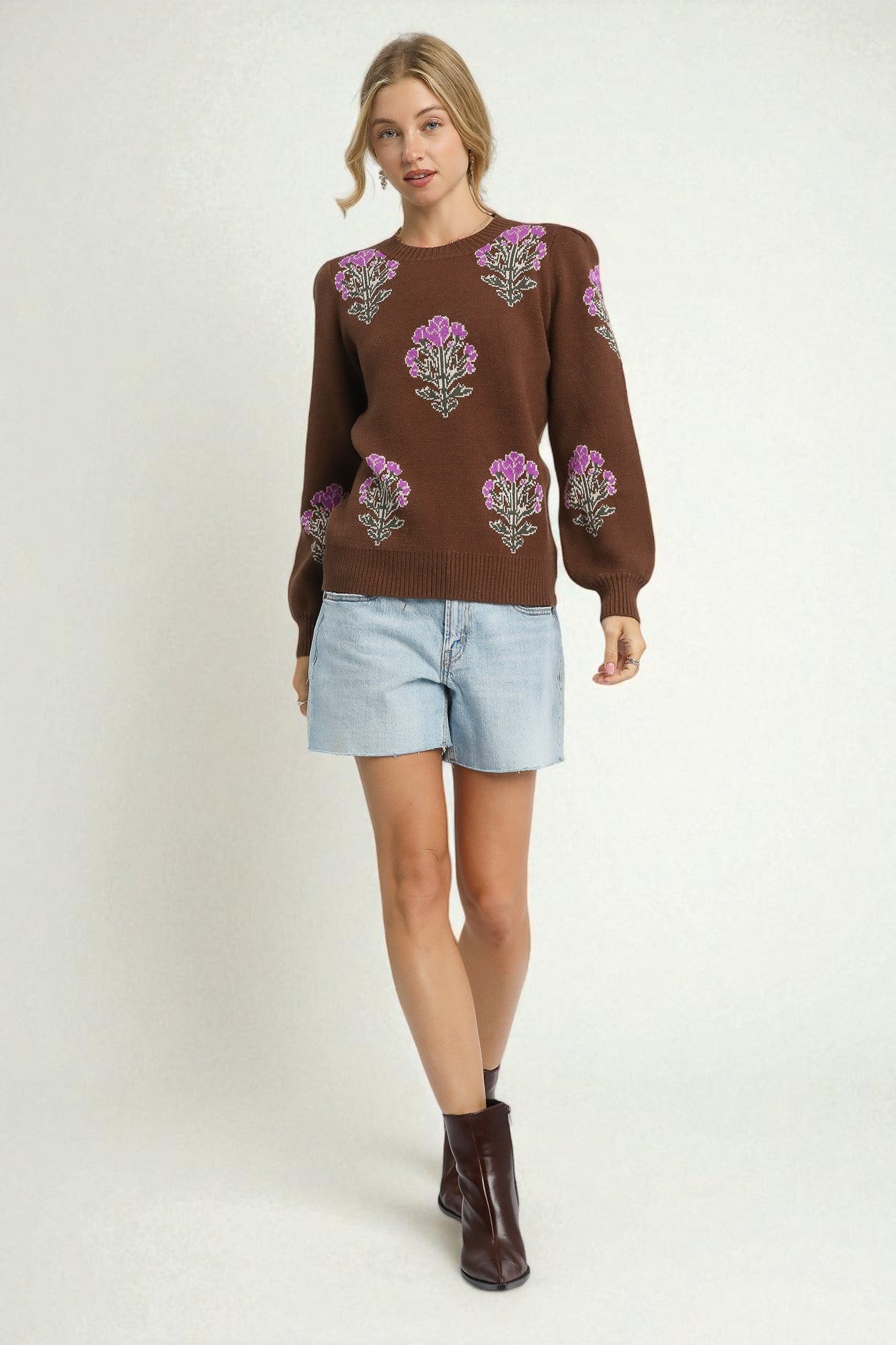 Woman wearing a brown sweater with floral patterns, light blue shorts, and brown boots on a white background